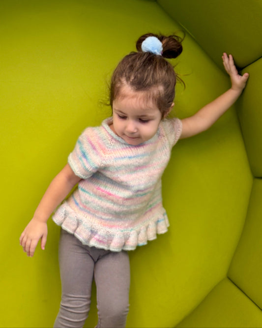 Child standing on a green cushioned chair wearing a colorful sweater and pink boots.