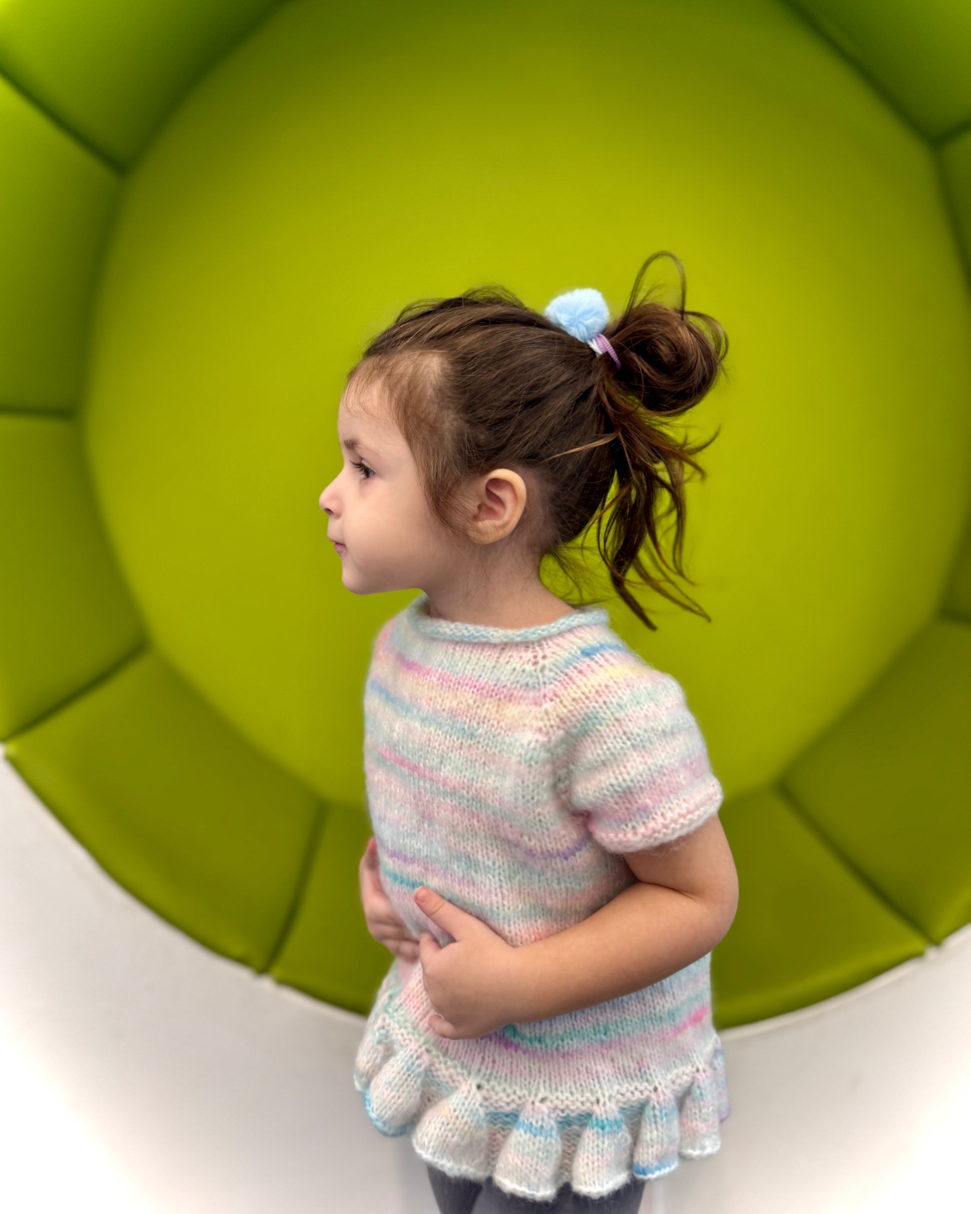 Young girl wearing a colorful knitted dress standing against a green wall.